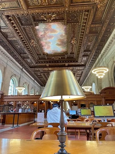 A photograph taken while sitting at a desk in the reference section of the main branch of the New York Public Library, showing a bronze lamp, beautiful wood shelves, floor-to-ceiling windows, and an ornately carved ceiling.