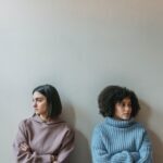 two young women stand against a wall their arms crossed at their chest facing away from one another.