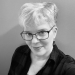 A greyscale close-up of me, standing in front of a blank background. I am a white woman with short silver hair cropped closely on the sides. I am wearing dark metallic rimmed glasses with rhinestones on the side. I’m wearing silver hook earrings with flat beads and a plaid shirt.