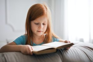 little girl with red hair reading a book on a sofa