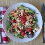 chicken and raspberry salad on a white plate on a bamboo mat with napkin and cutlery.