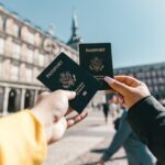 two hands each holding a blue US passports in the foreground against a background that looks like a European country.