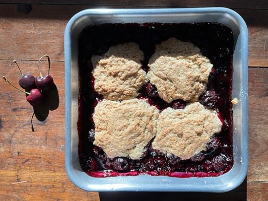 Baking pan with warm cherry cobbler and three fresh cherries next to it on a wooden table.