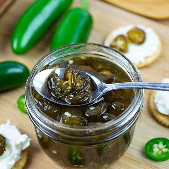A spoonful of Candied Jalapenos resting on the edge of a mason jar rim with crackers and cream cheese in the background.