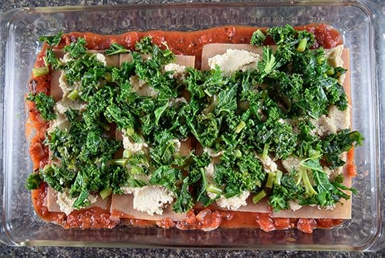 assembling strata in a casserole dish with layers of noodles, tomato sauce, cashew filling, and kale