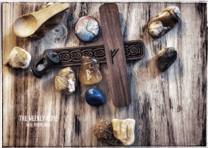 Runes cast on a medium dark wood grain background, with Fehu carved into a cedar stave, Berkana and Gebo carved into polished Baltic amber, a small wooden spoon, a tiny hourglass, a button with a treen in autumn, a piece of plished lapis lazuli, and other Baltic pieces scattered around, with the words 