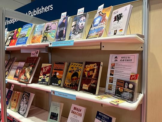 Novels by Adam Bender on a shelf at the ALA 2023 conference on a stand for independent publishers. Also displayed are bookmarks for Utopia PR and postcards for The Wanderer and the New West.