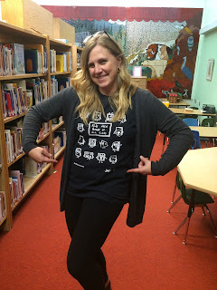 The author in her library. She has long blonde hair, is wearing all black and is pointing at her shirt, which says "Kids Need Books of All Kinds"