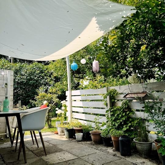 Eine Gartenszene, eine Terrasse, Tisch, Stuhl, Lampions, ein Sonnensegel darüber, ein sonniger Tag