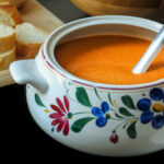 close-up of soup tureen filled with roast vegetable soup near a platter of bread.