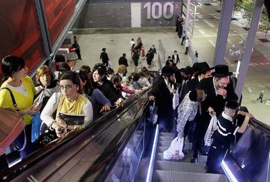 Women ride up one escalator while men in black suits and hats ride up a second escalator next to it, viewed from above.