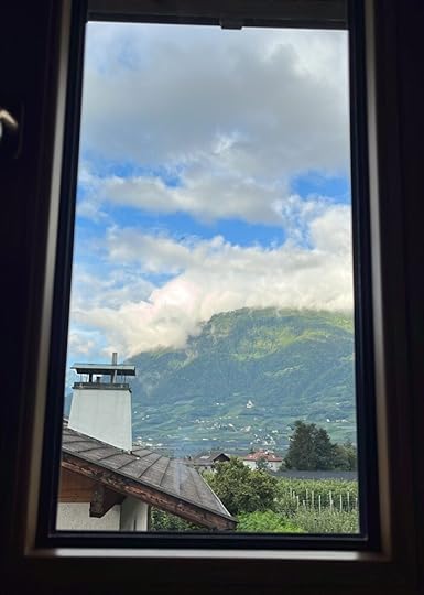 Blick durch ein schmales Fenster über ein Dach mit Schornstein auf Berge im Hintergrund, bewölkter Himmel, Weinstöcke in der Landschaft