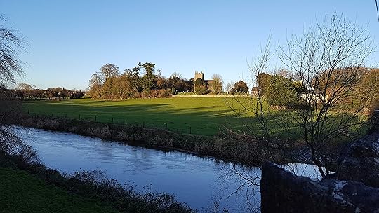 Church with tower, small in the distance with walled graveyard to right, surrounded by bare winter trees, beneath a cloudless blue sky, set in a rolling green landscape that sweeps down to a river in the foreground.
