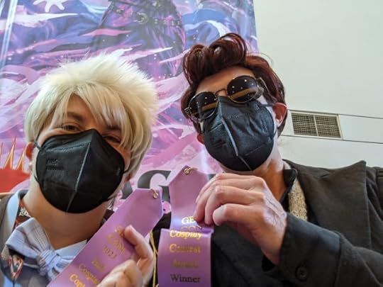 Two women in KN95 masks dressed as the angel Aziraphale, with a blong wig and tartan bow tie, and the demon Crowley, in a black blazer, red wig, and round black sunglasses, hold up purple award ribbons printed with the words