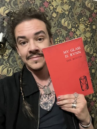 Photo shows Die Booth who is a middle aged white man with dark eyes, dark moustache, dark hair in plaits, holding up a copy of My Glass is Runn which has a red cover with a black woodcut print of an hourglass on it.