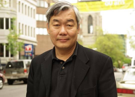 Color half portrait of Kwan, from mid-chest up. He's facing the camera, standing in front of a busy street with tall buildings across the street.