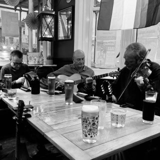 A bodhr��n player, guitarist and fiddler playing around a pub table with drinks on it.