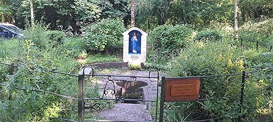 A circular space enclosed within a metal fence, exuberent flower beds in full growth but not yet in bloom, with steps in a gravel bed leading down to the water and beyond, a statue of Mary, to whom the well is dedicated. A wooden sign reads