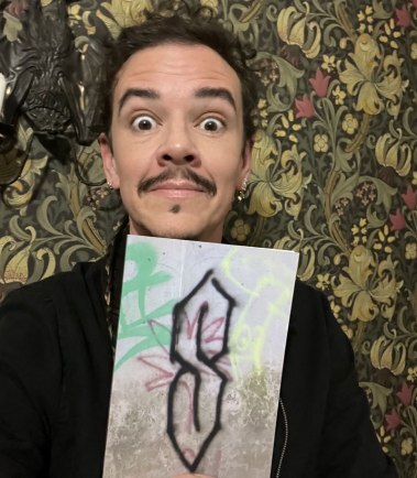 Image shows author Die Booth - a middle aged white man with dark hair, eyes and facial hair, pulling a wide-eyed face whilst holding up a copy of Cool S, which shows a graffiti Cool S drawn on a concrete wall on the cover.