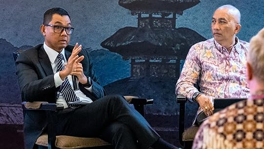 Picture of two men speaking at an event in front of a blue backdrop featuring traditional Balinese architecture. One man wears a suit and tie, the other a batik shirt.