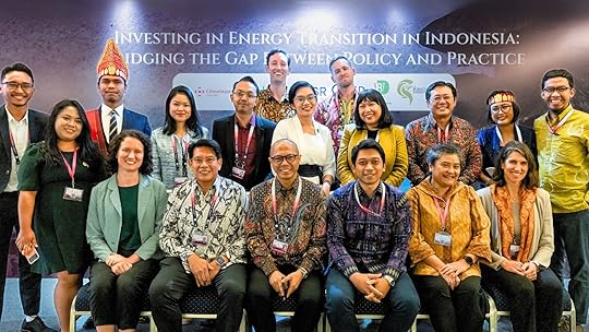A group photograph of men and women in formal western and Indonesian attire smiling at the camera. Behind them sits a backdrop reading