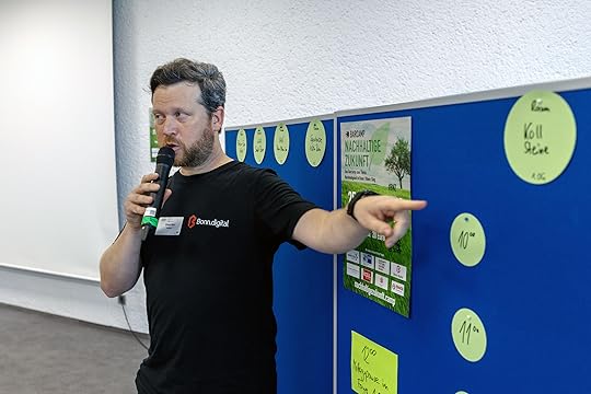 Ein abgekämpft aussehender Johannes spricht in ein Mikrofon, das er in der rechten Hand hält. Mit der linken zeigt er auf eine blaue Metaplanwand, auf der einige bunte Zettel hängen.