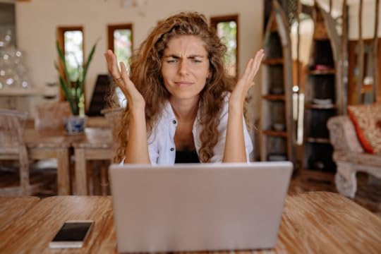 Image: a woman sits at a laptop computer with her hands in the air and a look of confused frustration on her face.