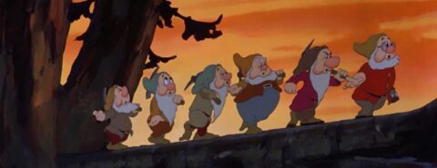 Screenshot of six of the seven dwarfs walking in a lie across a fallen tree log from the 1937 Disney film 
