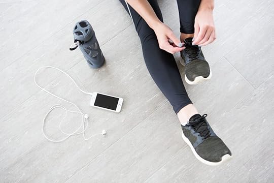 Woman sitting on hardwood floor lacing up her sneakers. Beside her is water bottle and smartphone with wired headphones.