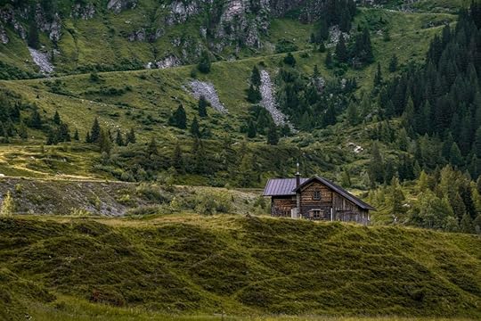 A wooden cabin in the hills, with a high mountainside covered in fir trees rising behind it.