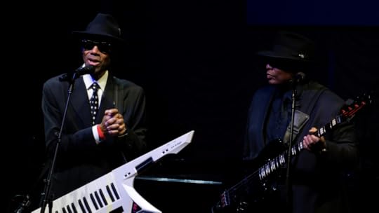 Two men in dark suits, hats and sunglasses standing side by side on a darkened stage. The one on the left holds a white keyboard, the one on the right holds a black bass guitar.