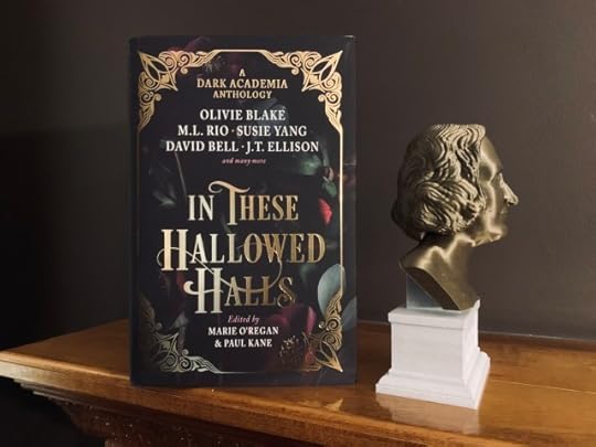 IN THESE HALLOWED HALLS (with cover art of gold foil and dark floral patterns) on a wood mantel next to the bronze-toned bust of Mary Shelley. The background is a dark plum-colored wall.