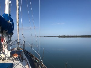 The prow of a yacht slices mirror-like waters reflecting at thin strip of land.
