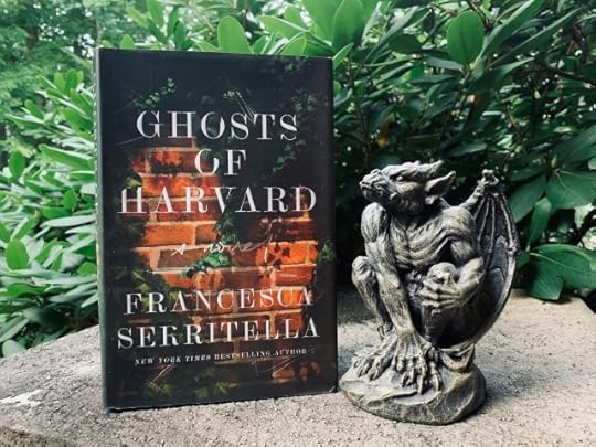 The image shows the novel THE GHOSTS OF HARVARD (with cover art of brick walls and ivy) sitting on an outdoor pedestal beside a stone gargoyle, with greenery in the background. Photo by AHS.