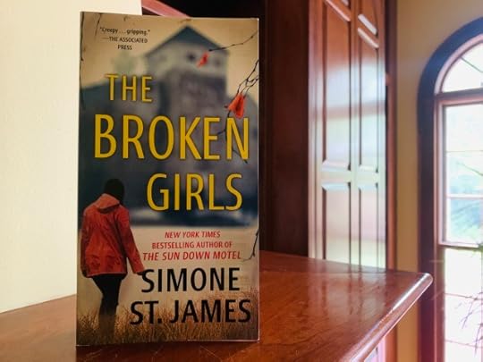 Shown is THE BROKEN GIRLS (with cover art depicting a young woman in a coat, beside a bare tree, approaching an out-of-focus building) on a wood mantel beside a window. Photo by AHS.