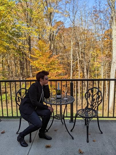 A woman dressed as Crowley from Good Omens, in a black jacket, black shirt, black jeans, black boots, a red wig, round black sunglasses, and a gray scarf necklace, sits at a wrought iron cafe table on a concrete deck. She has her elbow propped on the table and her chin in her hand, staring wistfully at the empty seat across from her. An unopened bottle of champagne and two empty champagne flutes sit on the table. The trees beyond the deck are festooned in yellow and orange autumn leaves.