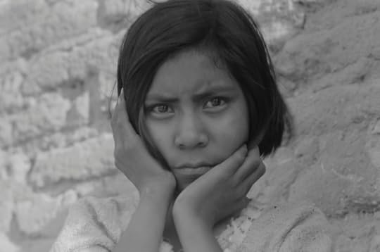 A pre-teen girl stares intentely at the camera, her hands cuppuibng her face. She stands in front of a light-colored brick wall, which sets off the darkness of her black hair.