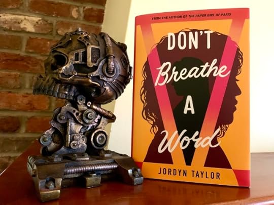 Pictured is DON'T BREATHE A WORD (with cover art depicting one figure in silhouette nested inside another) set on a mantel, before a brick wall, next to a steampunk skull in profile. Photo by AHS.