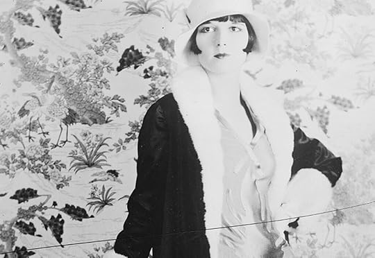 Half-length portrait of Louise Brooks, in a stylish coat and hat, one hand on a table.