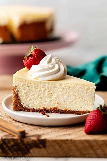 slice of cheesecake with whipped cream and strawberry on top on white plate on top of wood board.