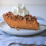 close up of slice of pumpkin pie with whipped cream on plate atop blue striped napkin.