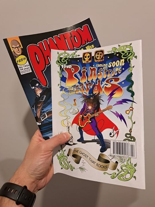Hand holding two copies of The Phantom #1956. the top copy shows the back cover, which reads: Coming soon: the Pinstripe Wizards. It shows the Phantom with a fake beard, a wizard hat, a cape and an electric guitar.