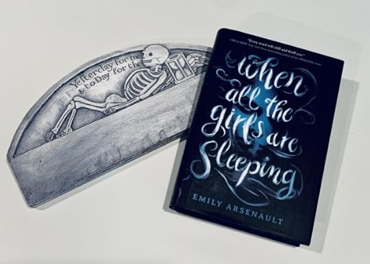 Shown is the book WHEN ALL THE GIRLS ARE SLEEPING (with cover art of a young woman's face in bluish-grey smoke) beside a mock headstone depicting a reclining skeleton with wings and an hourglass and the words, "Yesterday for me & toDay for thee." Photo by AHS.