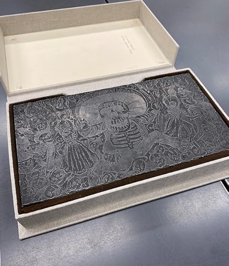 A large customized paper box is open to reveal a metallic block with the etching of a drawing of a menacing, machete-wielding, calavera surrounded by skeletons
