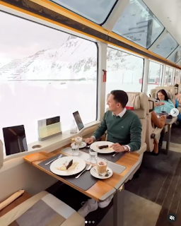 A man is sitting on a train, at a table. He's got a meal in front of him that looks as lovely as a restaurant, and the train window are huge. They go right to the carriage ceiling with the entire view being a snowy landscape.