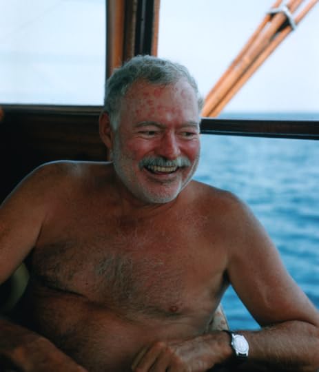 Ernest Hemingway, pictured from the waist up, at sea in the cabin of his boat, Pilar. The ocean is visible behind him. He is shirtless and laughing.