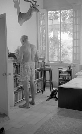 Ernest Hemingway, back to camera, stands at his bedroom bookcase, pecking at the typewriter on top of it. he is shirtless, wearing swimming trunks and sandals. A stuffed gazelle head is mounted on the wall above him. The large window behind him is open, showing a tall tree and bright sunshine outside.
