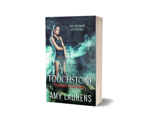 Amy Laurens' novel Touchstone: a woman in tank top and shorts and fingerless gloves stands near an industrial building in low lighting, with green mist trailing around her. The tagline reads, Just one touch will kill you. The series title, Shards Of Fate, stands out in orange below the book's title.