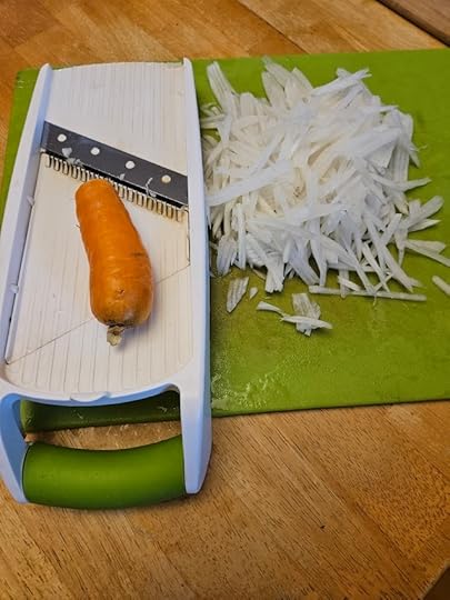 Cutting board with shredded radish on one side and a carrot sitting on top of a mandolin ready to be grated as well.