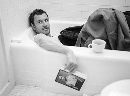 Michael Fassbender reclines clothed in a bathtub, holding a copy of a Genet book, for T Magazine